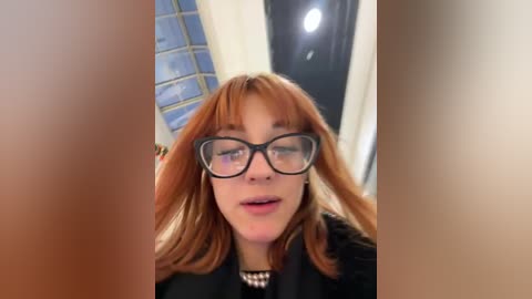 Media: Video of a Caucasian woman with fair skin, straight auburn hair, and glasses, wearing a black coat and standing in a modern room with blue-tiled walls and a door.