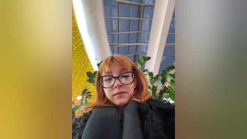 Media: A video of a fair-skinned woman with red hair and glasses, wearing a dark coat, sitting in a modern indoor space with green plants and a large glass ceiling.