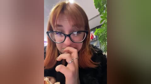 Media: Video of a young woman with fair skin and reddish-brown hair, wearing glasses and a black fur coat, eating a sandwich, with greenery in the background.