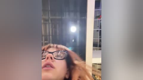 Media: Video of a woman with glasses, fair skin, and light brown hair, seen from a side angle. Background shows a dimly lit room with shelves and a bright light source.