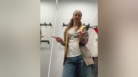 Media: Video of a blonde woman in a changing room, wearing a white T-shirt, jeans, and a jacket, with a casual expression, surrounded by clothes on racks.