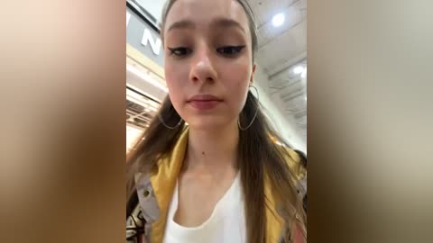 Media: A video of a young woman with fair skin, brown hair in pigtails, wearing a yellow and black floral jacket over a white top, standing in a mall with a blurred background.