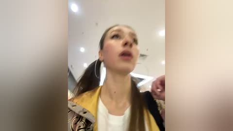 Media: Video of a woman with long brown hair, wearing a yellow and black jacket, looking surprised in a mirrored hallway.