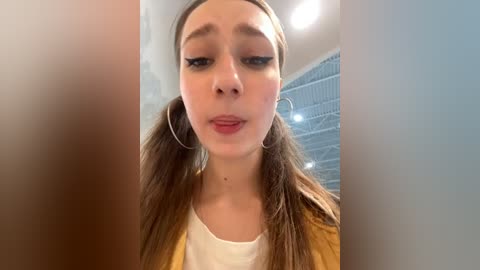 Media: Video of a young woman with long, straight brown hair styled in pigtails, wearing hoop earrings and a yellow jacket over a white shirt, indoors with a blurred industrial background.