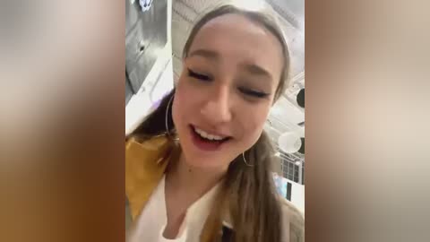 Media: Video of a young Asian woman with light skin and straight, light brown hair, wearing a white headband and a yellow jacket, smiling widely, taken from a selfie angle.