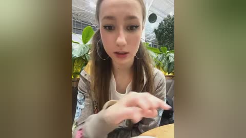 Media: Video of a young Caucasian woman with light skin and brown hair in pigtails, wearing a light-colored jacket, seated in a greenhouse, holding a plant.