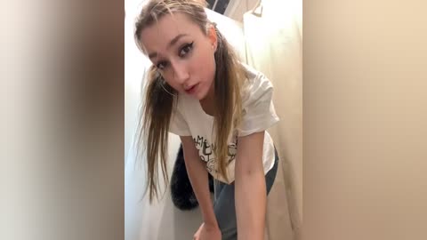 Media: Video of a young Caucasian woman with straight, light brown hair in pigtails, wearing a white t-shirt and gray shorts, leaning forward, looking into a narrow hallway.