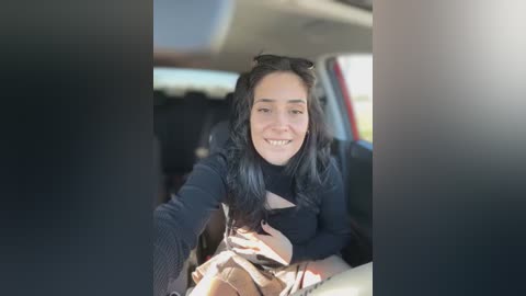 Media: Video of a smiling Latina woman with long black hair, wearing a black sweater, seated in a car with a blurred background.