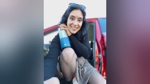 Media: Video of a smiling woman with dark hair, wearing a black sweater and grey skirt, sitting in a red car, holding a blue and green water bottle.