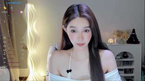 Media: Video of a young Asian woman with long, straight black hair, fair skin, and a slender physique. She wears a white off-shoulder top. Background includes a wall, a white shelf with decorative items, and a lamp.