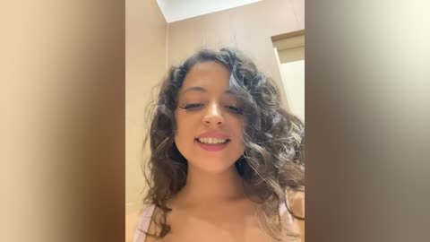 Media: Video of a young woman with curly, shoulder-length brown hair, light skin, and a warm smile. She wears a pink bra, standing indoors against a beige wall.