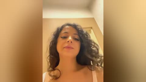 Media: Video of a young woman with curly dark hair, wearing a white top, standing in a dimly lit room with beige walls and a doorway visible in the background.