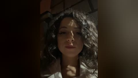 Media: Video of a young woman with curly black hair, fair skin, and a neutral expression, illuminated by dim light. She wears a white blouse, suggesting a casual setting.