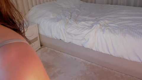 Media: A video of a disheveled, unmade bed with white sheets, partially covered by a beige blanket. The image captures a close-up view of a person's arm and shoulder in the lower left corner, suggesting a bedroom setting.