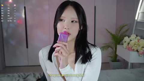 Media: Video of an East Asian woman with straight black hair, wearing a white top, eating a purple lollipop. Background features a pastel-toned room with white furniture and potted plants.