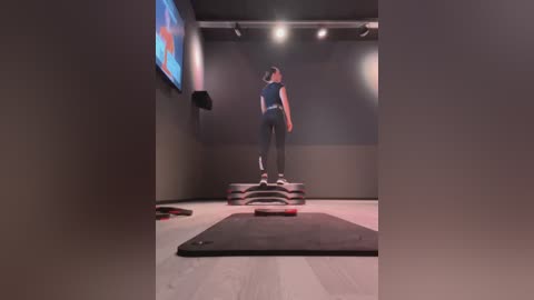 Media: Video of a woman in a dark sports bra and black leggings standing on a balance board in a dimly lit, minimalist gym.