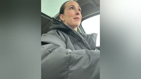 Media: A video of a woman in a gray, puffy winter coat, sitting in a car. She has her hair tied back, and her expression is contemplative, looking out the window. The car interior is dimly lit.