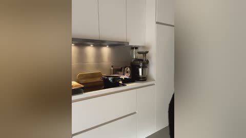 Media: Video of a modern, minimalist kitchen with white cabinets, stainless steel appliances, a black stovetop, and a beige countertop. A small round table with a yellow cloth is visible. The lighting is soft, highlighting the sleek, clean design.