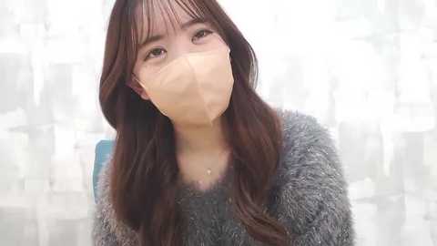 Media: Video of an Asian woman with long brown hair, wearing a face mask and a gray fuzzy sweater, in a softly blurred white background.