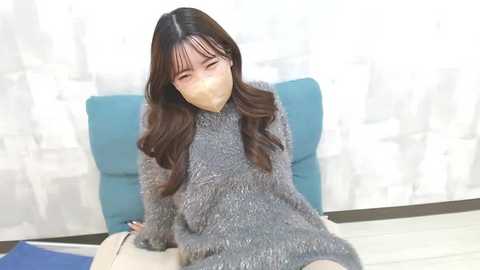 Media: Video of a young Asian woman with long, straight brown hair, wearing a grey knit sweater, sitting on a blue cushion, with a white mask covering her mouth and nose, in a minimalist room with white curtains.