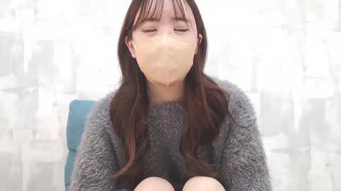 Media: Video of an East Asian woman with long, straight dark hair, wearing a gray fuzzy sweater and a beige surgical mask, sitting in a chair against a white, semi-transparent curtain backdrop.