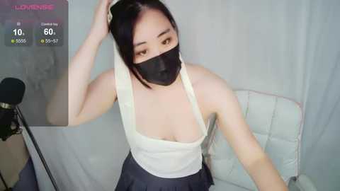 Media: Video of an Asian woman with long black hair, wearing a white tank top and black skirt, seated on a white couch. She has a black face mask, with a digital clock overlay showing 60% humidity.