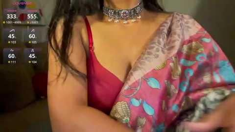 Media: Video of a woman with medium skin tone, wearing a red bra and a colorful, patterned sari, adorned with a floral necklace. The background shows a dimly lit room with a TV screen displaying live stream details.