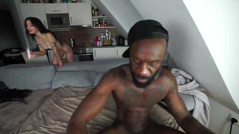 Media: A video captures a nude, muscular Black man with a tattooed chest sitting on a bed in a modern, minimalist bedroom. A mirror behind him reflects a topless woman with long hair.