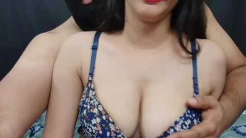 Media: Video of a woman with medium skin tone, wearing a blue floral bra, revealing ample cleavage. A man's arm is around her neck, partially visible.