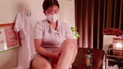 Media: Video of an Asian woman in a white mask and uniform, sitting on a massage table with a potted plant, in a dimly lit room with posters on the wall.