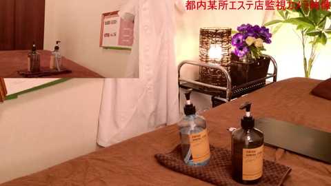 Media: Video of a spa room with a white robe hanging, a wooden table with lotion bottles, a vase of purple flowers, and a candle-lit wicker basket.