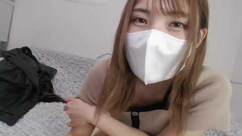 Media: Video of an Asian woman with long brown hair, wearing a white mask, beige top, and gray sweater. She sits on a bed with a gray blanket, in a simple, minimally decorated room.