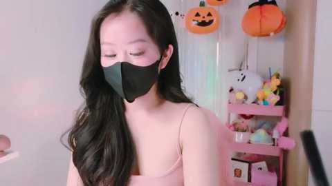 Media: Video of an Asian woman with long black hair, wearing a black face mask and pink spaghetti strap top, in a Halloween-themed room with orange pumpkin decorations.