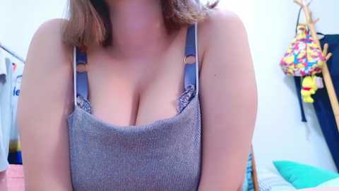 Media: Video of a light-skinned woman with medium-length brown hair, wearing a grey sleeveless top with blue straps, revealing cleavage, standing in a brightly lit bedroom with colorful bags hanging on the wall.