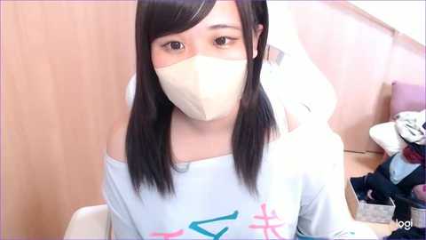 Media: A video of an East Asian woman with long black hair and a white mask, wearing a light blue off-shoulder top with a colorful graphic, seated indoors.