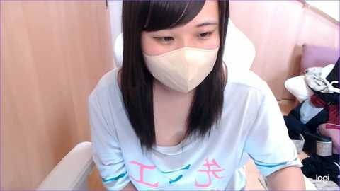 Media: Video of an East Asian woman with long, straight black hair, wearing a white T-shirt with Japanese characters, a beige mask, and a white surgical cap, sitting in a wooden chair in a cluttered room.