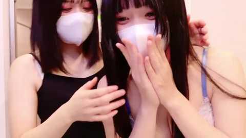 Media: A video of two young Asian women with long black hair, wearing white face masks, black tank tops, and holding each other's hands. The background is a plain, light-colored room.