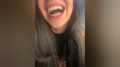 Media: Video of a smiling woman with long, dark hair, wearing a black top and silver necklace, set against a blurry, beige background.