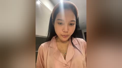 Media: Video of an Asian woman with medium skin tone, straight black hair, and a pink headband, wearing a light pink button-up shirt, indoors with blurred background.