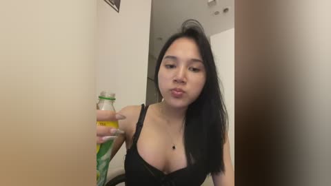 Media: A video of a young Asian woman with long black hair, wearing a black lace bra, holding a green and yellow drink bottle, indoors against a beige and brown wall.