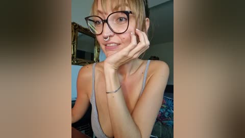 Media: Video of a slender, blonde woman with a septum piercing, wearing glasses and a low-cut tank top, posing in a dimly lit room with a mirror and colorful blankets in the background.