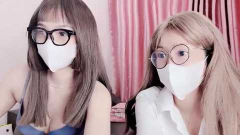 Media: Video of two East Asian women with long hair, wearing black-framed glasses, white face masks, and light-colored shirts, seated in front of pink curtains.