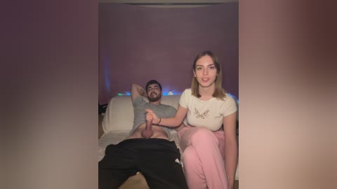 Media: Video of a bearded man in a grey t-shirt, seated on a bed with a woman in a white top and pink pants, both looking at the camera.