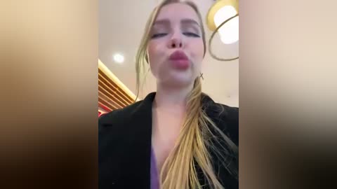 Media: A video of a blonde woman with light skin, wearing a black blazer and a purple top, blowing a kiss. Background features modern lighting and wooden paneling.