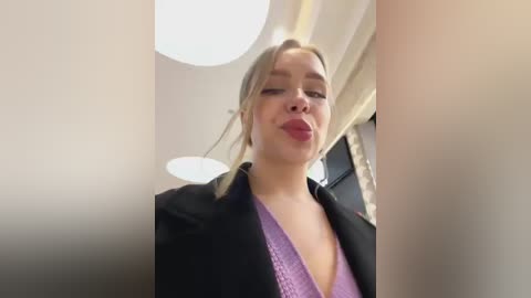 Media: Video of a young, fair-skinned woman with blonde hair in a ponytail, wearing a black blazer over a purple lace top, kissing her reflection in a mirror. Background shows modern, light-colored decor with a circular light fixture.