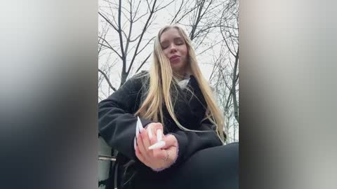 Media: Video of a blonde woman with long hair, wearing a black jacket, sitting on a black car seat, holding a white cigarette, surrounded by bare trees in an overcast outdoor setting.