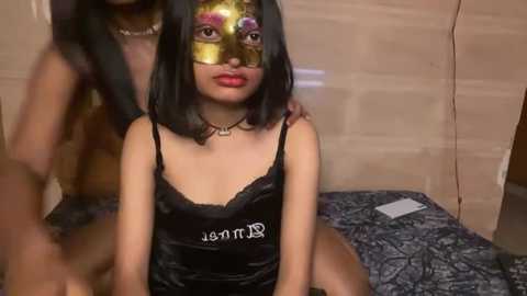 Media: Video of a young woman with medium brown skin and straight black hair, wearing a black satin camisole, gold Venetian mask, and a necklace. She sits on a bed with patterned sheets in a dimly lit room.