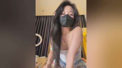 Media: Video of an Asian woman with long black hair, wearing a black face mask, a white strapless top, and a yellow jacket, sitting on a bed with beige walls and a circular mirror in the background.