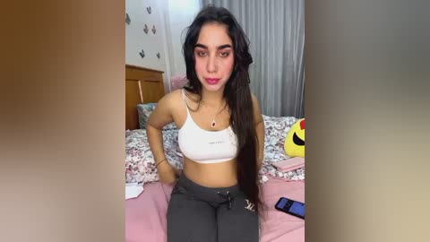 Media: Video of a young South Asian woman with long, dark hair, wearing a white crop top and black pants, sitting on a bed with floral sheets in a cozy bedroom.