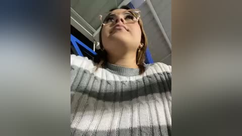 Media: Video of a young woman with light skin, wearing glasses, and a grey and white striped sweater, looking upward with a contemplative expression. The background features a blue and grey office setting.